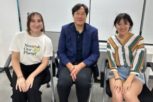 Two women sit on each side of a man in a blue blazer, all smiling at the camera. The women wear casual summer clothing, and the room has large windows in the background.