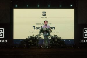 A young man stands on stage behind a podium decorated with flowers, delivering a speech in front of a large screen displaying the KKOOM logo and the title “Dream Big Speech: Taehyeon.” The stage is dimly lit with spotlights above.