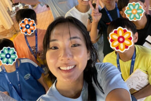A volunteer takes a cheerful selfie with a group of children standing behind her. The children’s faces are covered with flower stickers, and they are smiling and making peace signs.