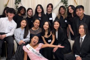 A large group of young adults, dressed in semi-formal black and white attire with name tags, pose together for a cheerful group photo at an indoor event. One woman in the front playfully extends her arms while wearing a sash that reads “Miss Universe.” Everyone is smiling and gathered closely, creating a warm, lively atmosphere.