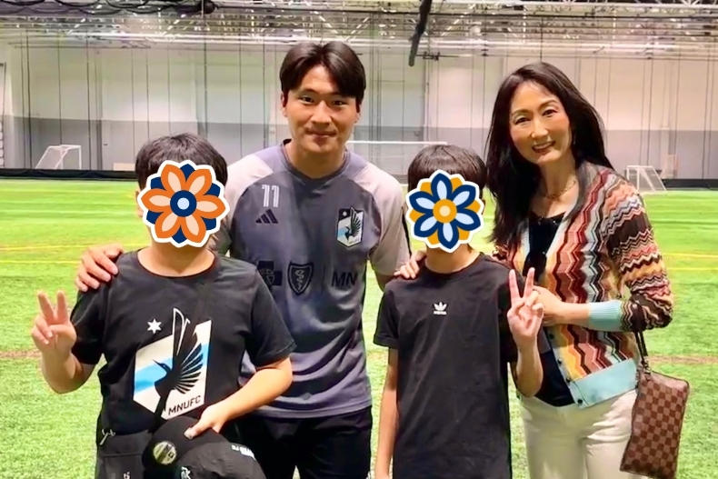 Two boys — their faces covered with floral stickers — stand on a turf soccer field (indoor facility) alongside a male soccer player in his team jersey and Patty (or a woman) standing to the side, smiling. The boys hold up peace signs. The background shows a large indoor field with netting and white walls. The lighting is bright and the mood is excited and proud.