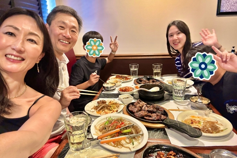 A cheerful group of two adults (a woman and a man) and two boys sit together at a restaurant table filled with Korean dishes — plates of grilled meat, japchae, side dishes, glasses of water, and chopsticks poised to eat. The adults smile at the camera; the boys’ faces are covered by colorful flower stickers. The setting is cozy and casual, giving a warm, family-style vibe.
