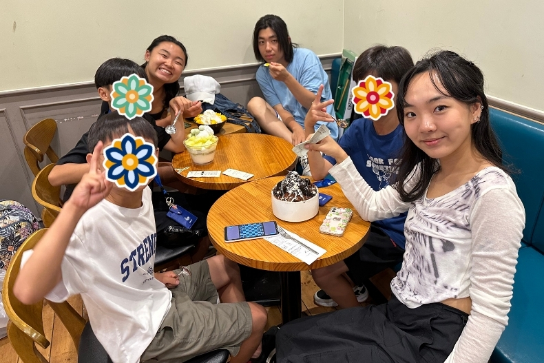 A group of volunteers and teenagers sits around a wooden café table enjoying drinks and desserts. They smile and make peace signs toward the camera. Several colorful sticker icons cover their faces. The café setting includes small plates, cups, and a cozy indoor atmosphere.