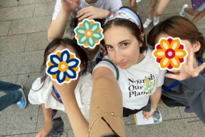 A group of girls pose playfully for a selfie with a volunteer. Their faces are covered with stickers as they smile and gesture toward the camera.