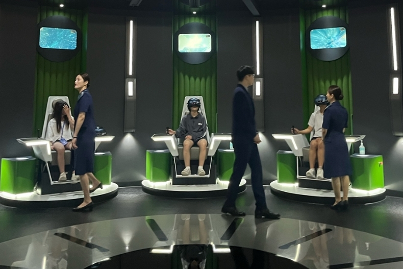 Girls experience a virtual reality exhibit, each seated in futuristic green chairs wearing VR headsets as staff guide the activity. Faces are covered.