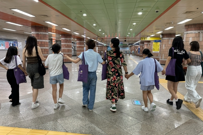 A group of girls and volunteers walk hand-in-hand through a subway station, carrying matching purple tote bags.