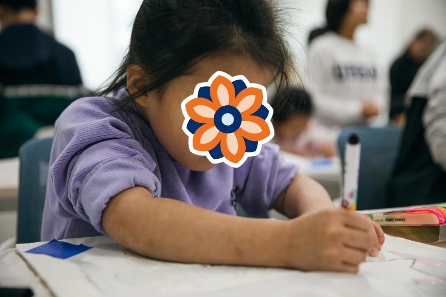 A young child sits at a desk and focuses on drawing or writing during an activity. Their face is covered with a sticker.