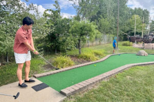 A young person plays mini-golf on an outdoor course surrounded by greenery. Another player is seen in the distance.