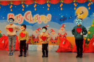 Four children wearing sweaters with heart designs stand on stage in front of a colorful Christmas-themed backdrop. Their faces are covered with flower stickers, and holiday decorations fill the background.