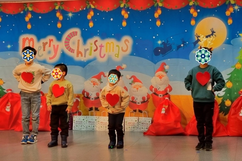 Four children wearing sweaters with heart designs stand on stage in front of a colorful Christmas-themed backdrop. Their faces are covered with flower stickers, and holiday decorations fill the background.