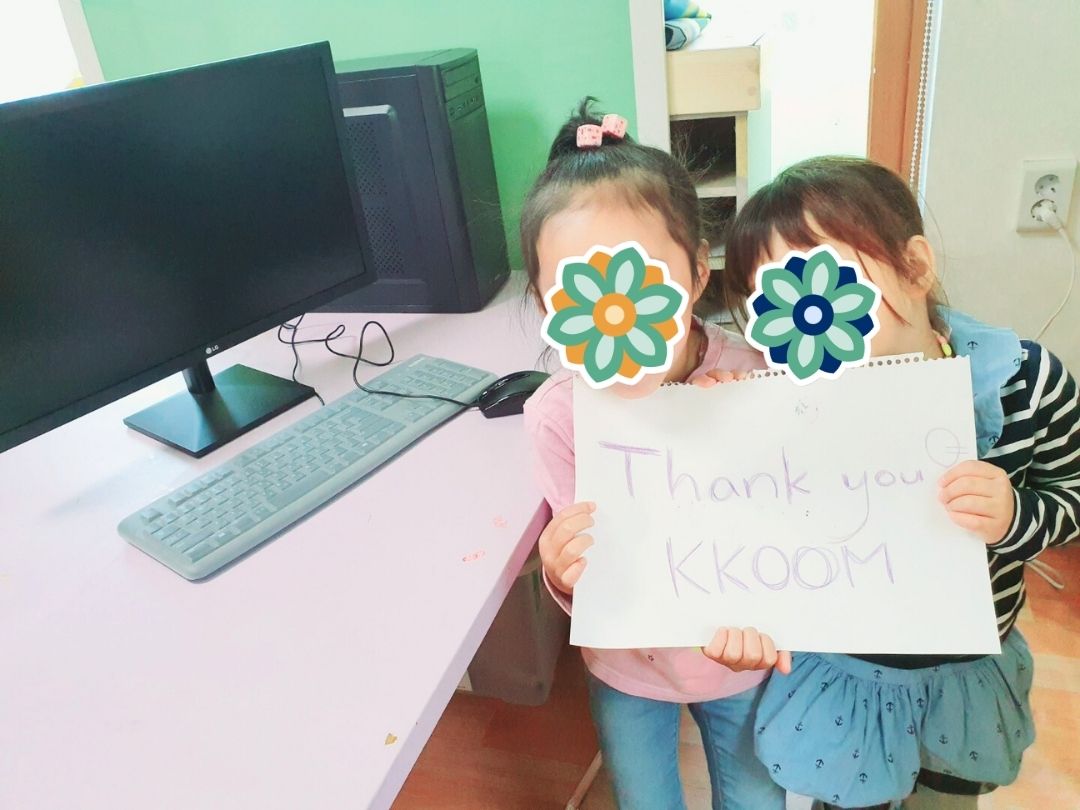 Two children holding a sign that says ‘Thank you KKOOM’ next to their new computers.