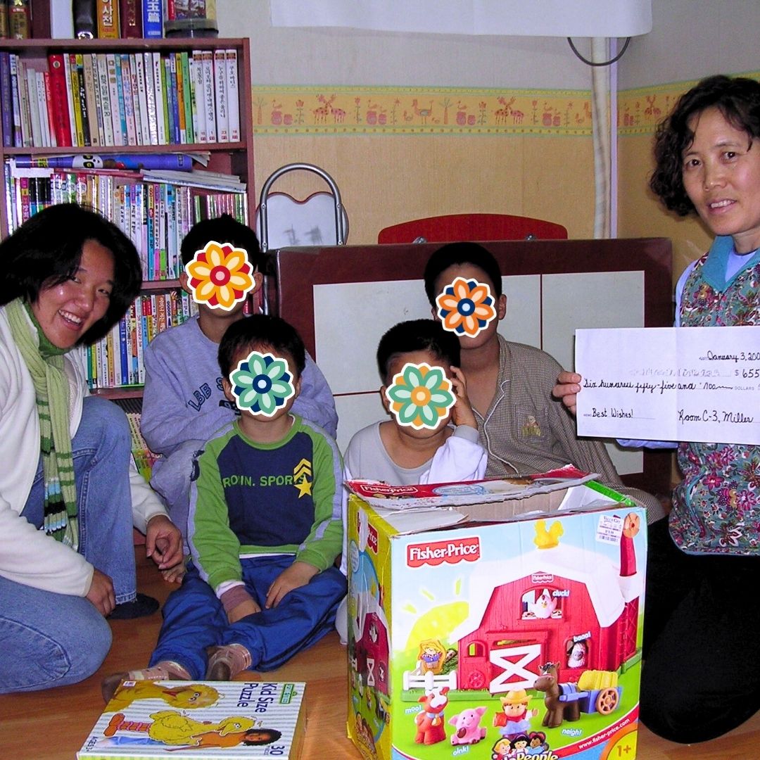 House mom holding a large donated check while sitting with the children and KKOOM’s co-founder during a holiday event.