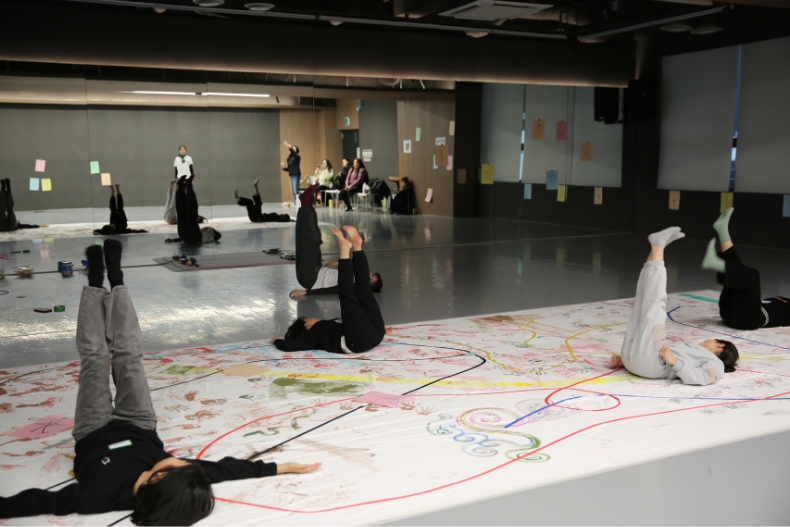 Students participate in a movement activity in a large studio, with drawings spread out on paper across the floor.