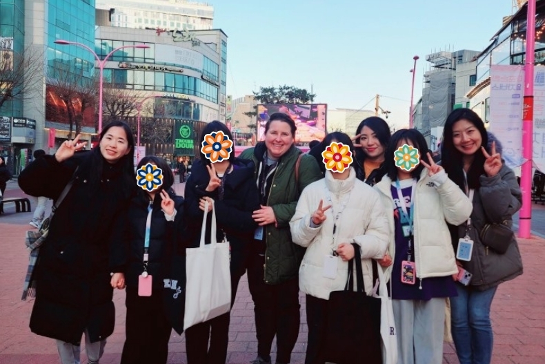 A group of volunteers and girls stand together outdoors in a city plaza, smiling and making peace signs. The girls’ faces are covered with flower stickers.