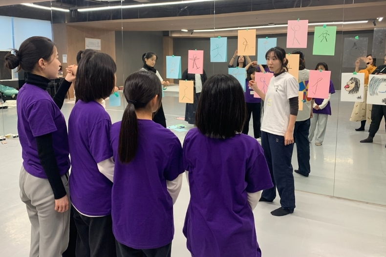 Girls wearing purple shirts stand in a dance studio facing an instructor, with colorful papers displayed on the mirrors in front of them.Girls wearing purple shirts stand in a dance studio facing an instructor, with colorful papers displayed on the mirrors in front of them.