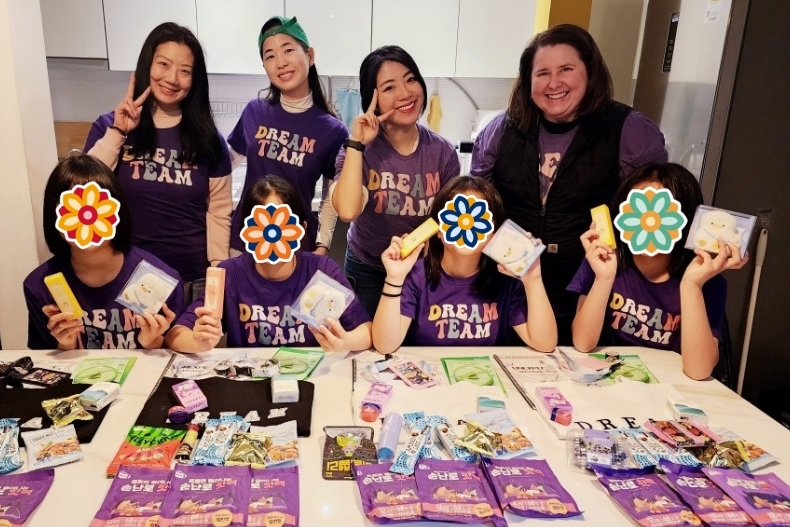 A volunteer and several girls wearing purple shirts sit at a table covered with colorful crafts and materials. The girls’ faces are covered with flower stickers.