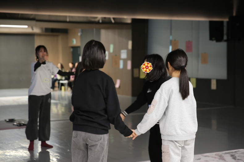Four girls hold hands while practicing a movement activity in a large studio space. Their backs face the camera.