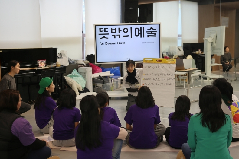 A group of girls and volunteers sit on the floor in a studio listening to an instructor at the front.