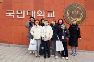 A group of volunteers and girls stand together smiling in front of a large wall with the Kookmin University sign and emblem. The girls’ faces are covered with flower stickers, and several hold tote bags labeled “DREAM.” The group is bundled in winter coats.
