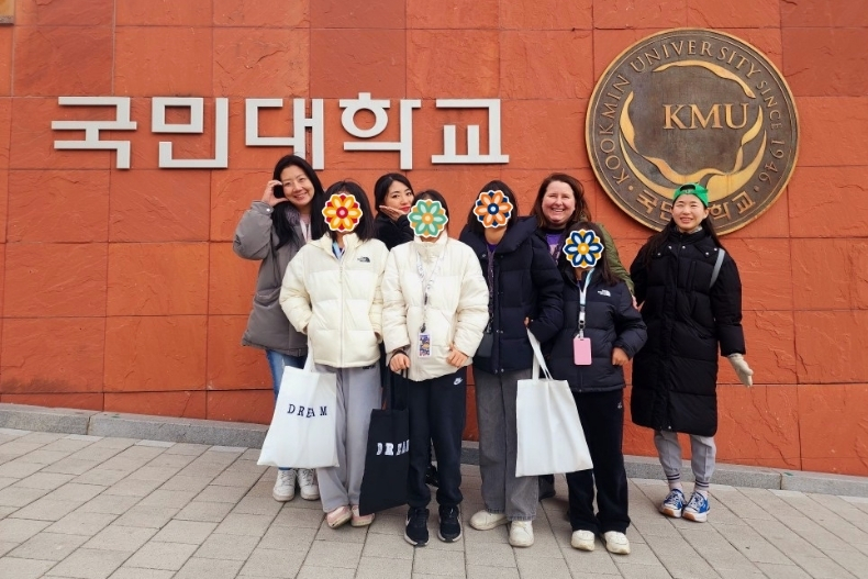 A group of volunteers and girls stand together smiling in front of a large wall with the Kookmin University sign and emblem. The girls’ faces are covered with flower stickers, and several hold tote bags labeled “DREAM.” The group is bundled in winter coats.