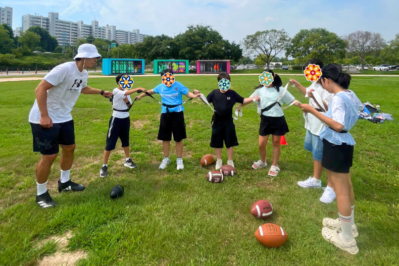 Children participating in an outdoor sports activity on a grassy field, with faces covered for privacy.