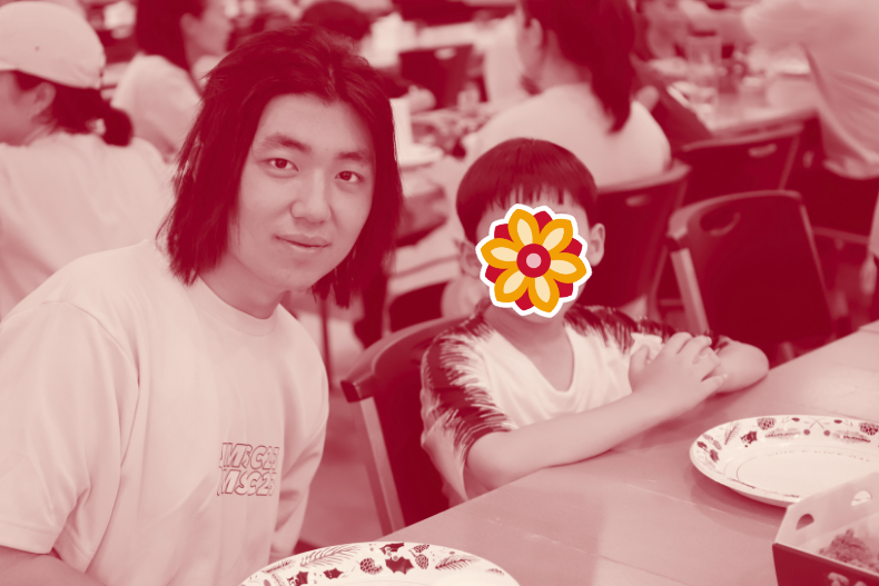 A young man sitting at a table with a child whose face is covered with a flower sticker. They appear to be in a classroom or activity setting.