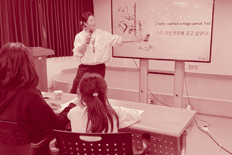 A classroom scene with an adult presenting at the front of the room beside a screen while children sit at desks facing forward.