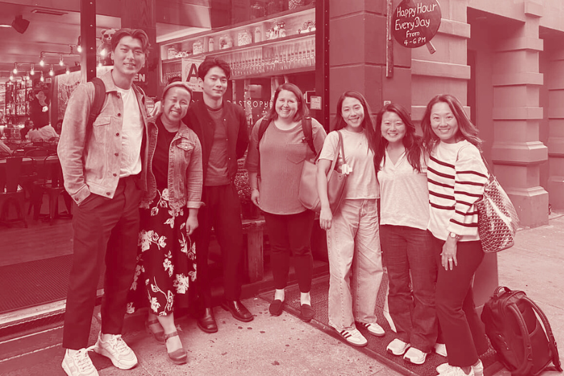 A group of seven adults standing together outside a storefront at night, smiling for a group photo.A group of seven adults standing together outside a storefront at night, smiling for a group photo.