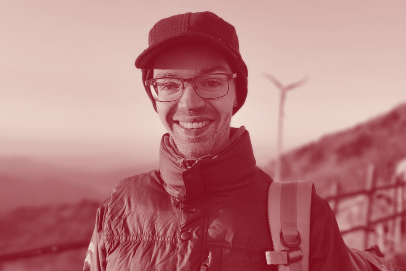 A man wearing glasses and a cap smiling outdoors with mountains and wind turbines in the background.
