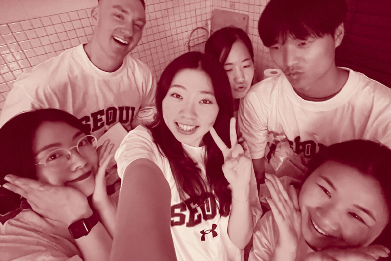 A group of five young adults in matching shirts gathered closely for a playful indoor selfie.