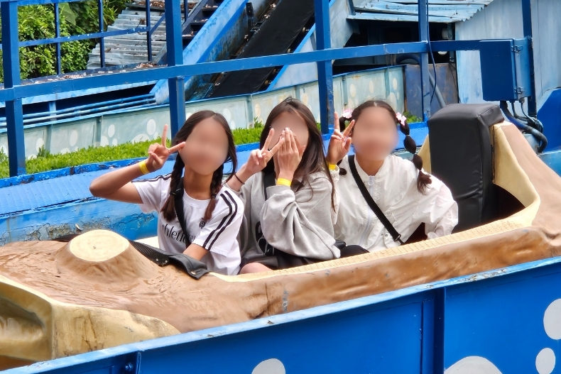 Three girls riding a water log flume attraction at an amusement park, smiling and posing with peace signs.