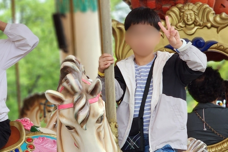A boy riding a carousel horse at an amusement park, waving a peace sign at the camera.