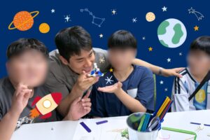 A KKOOM volunteer and students pose playfully together at a STEAM and English workshop, surrounded by illustrated planets, stars, and a rocket on a night-sky background.