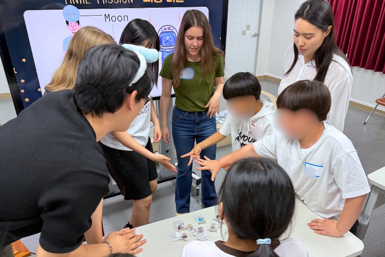 Students and volunteers play rock, paper, scissors around a table to decide prize order after the final mission at the STEAM workshop.