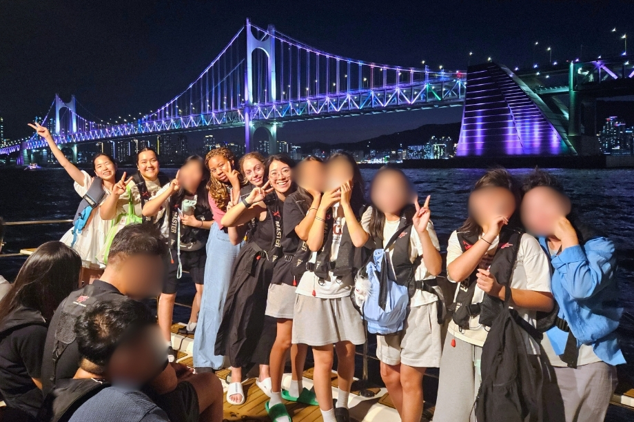 Students and volunteers on a sunset yacht cruise along the Busan coastline, with the Gwangan Bridge lit up in the background.