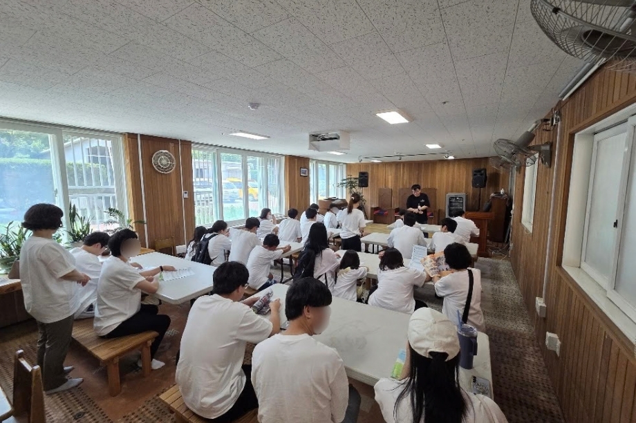 Picture of a big classroom full of kids and volunteers sitting at tables together listening to the volunteer leader.